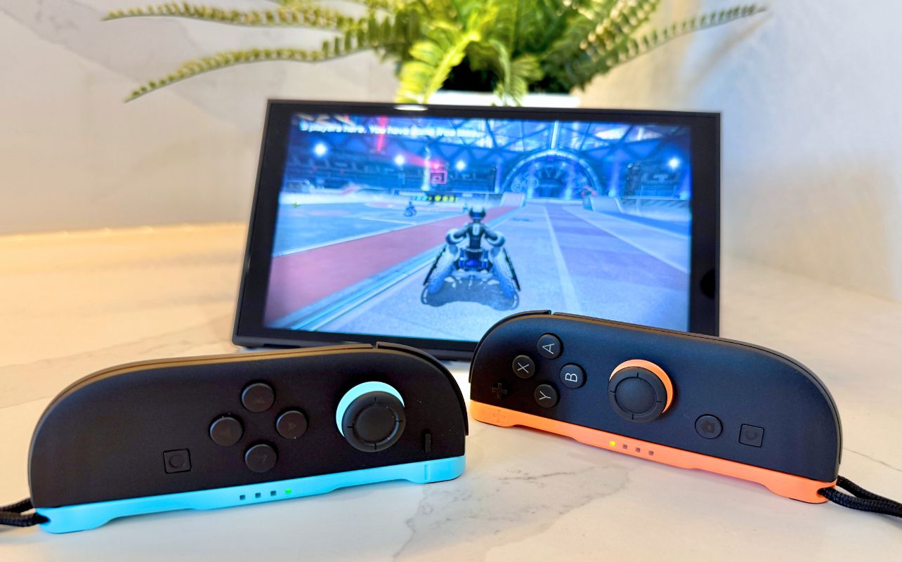 Two Joy-Con 2 gaming controllers, one blue and one orange, are in front of a Switch 2 tablet displaying Drag x Drive. A plant decorates the background.
