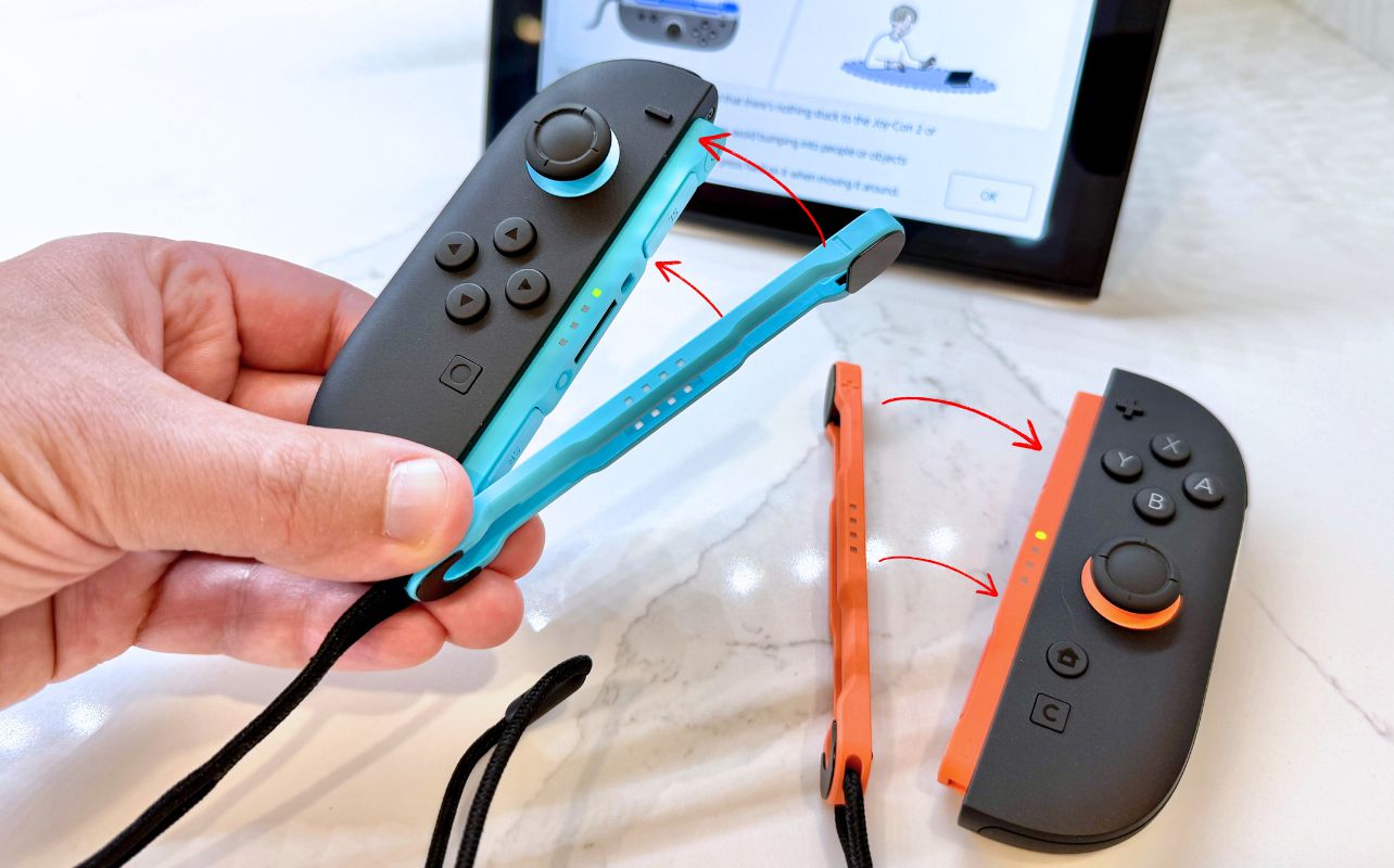 Hand holding a blue and black game controller, with another orange controller and a strap on a marble surface. The tablet in the background shows instructions.