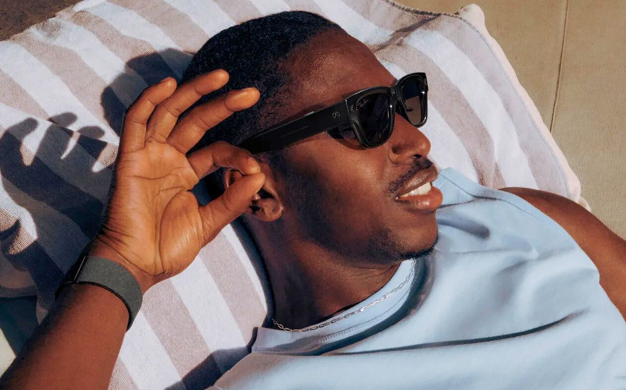 A man lays in the sun wearing the Meta Ray-Ban Display and Neural Band, moving his hand to select