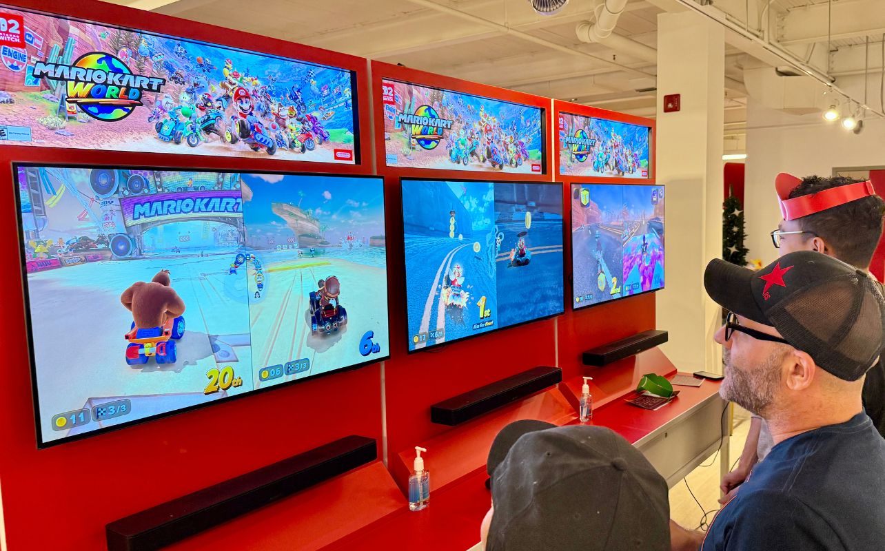 Three people watch a "Mario Kart World" video game on four screens in a brightly lit room. Game action shows karts racing. Feeling focused.