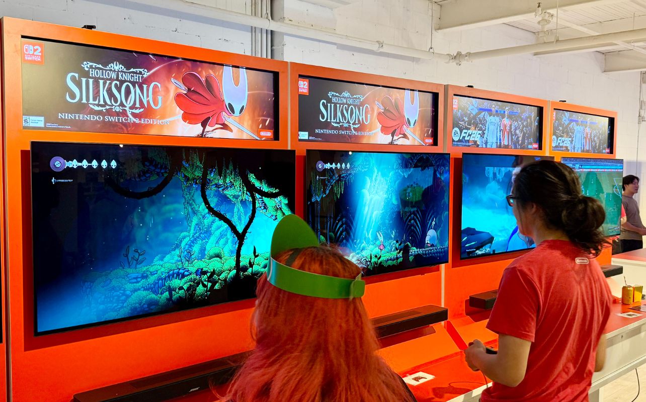 People playing "Hollow Knight: Silksong" on Nintendo Switch 2 at a gaming booth. Bright displays, red decor, focused mood.