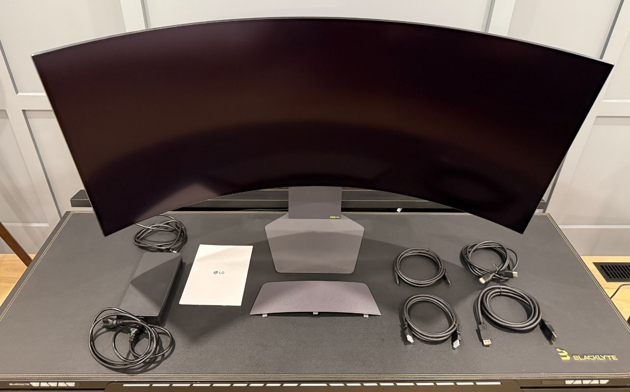 Curved LG UltraGear 5K2K monitor on a desk with power adapter, cables, and a manual.
