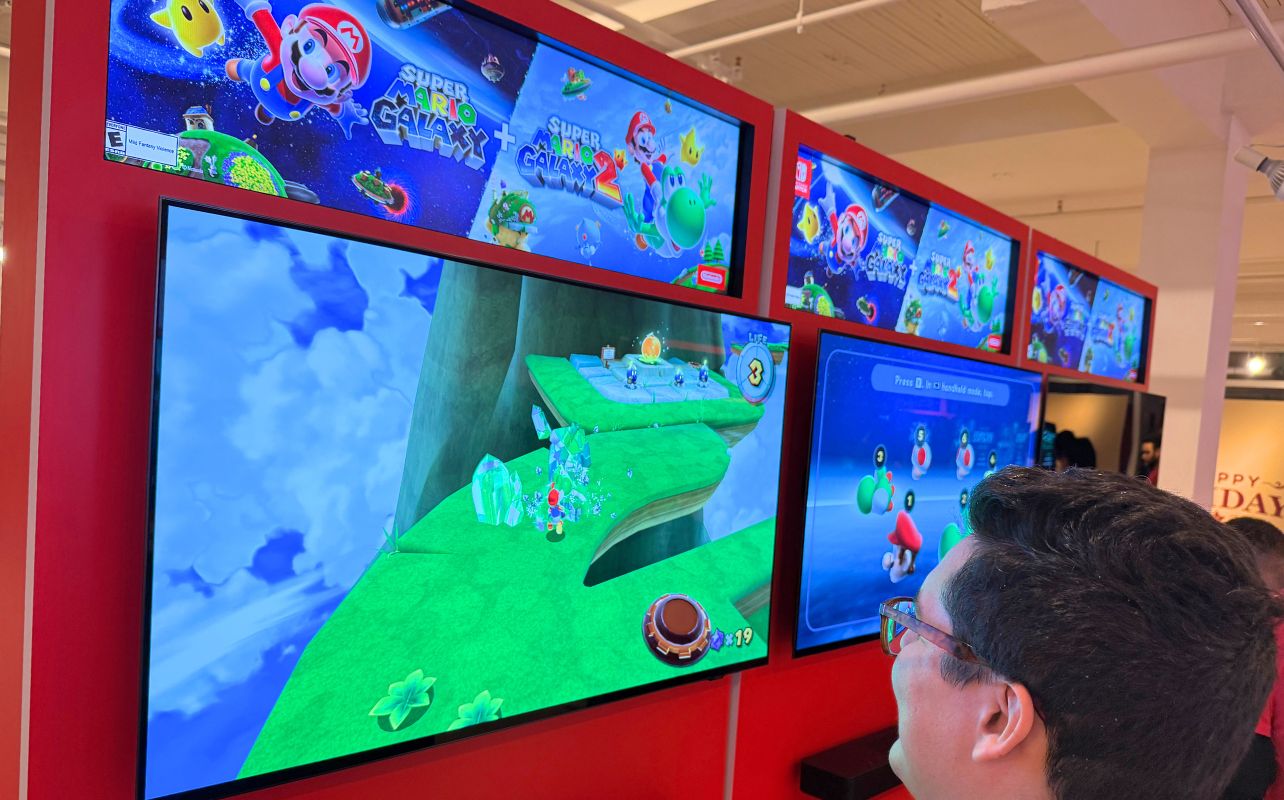 Person plays Super Mario Galaxy on a large screen in a gaming expo. Bright colors, red display stand, game logos visible above.