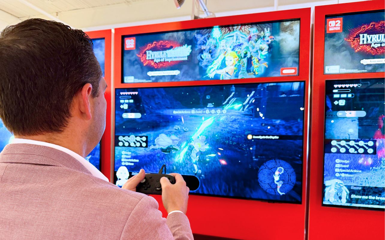 Matthew Rondina in a pink jacket plays a video game on a screen with a "Hyrule Warriors: Age of Calamity" display, set in a bright red booth.
