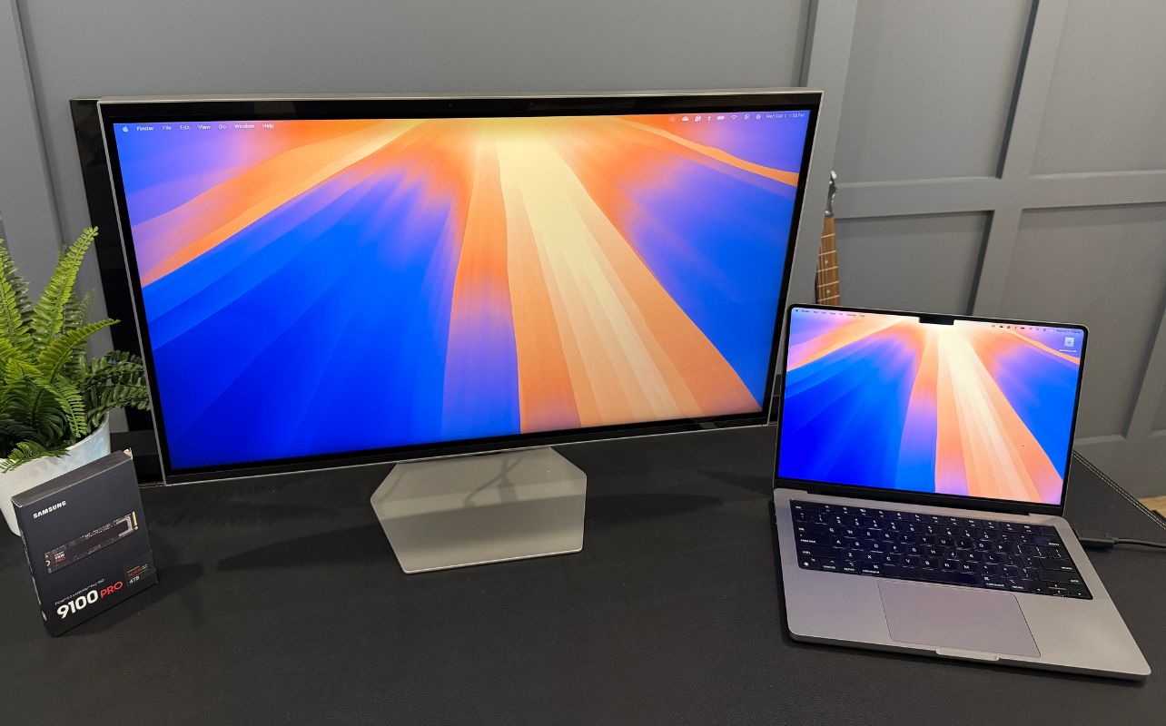 Samsung Odyssey 3D monitor and a laptop display orange and blue abstract designs on a desk. Beside them is the Samsung 9100 Pro box.