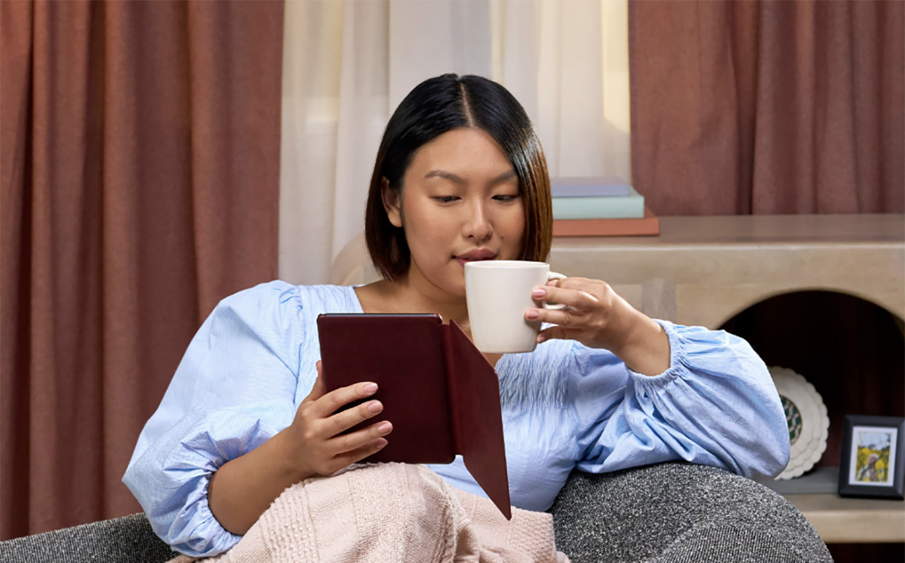 A woman in a chair with a coffee mug in one hand and an Amazon Kindle Colorsoft in the other.