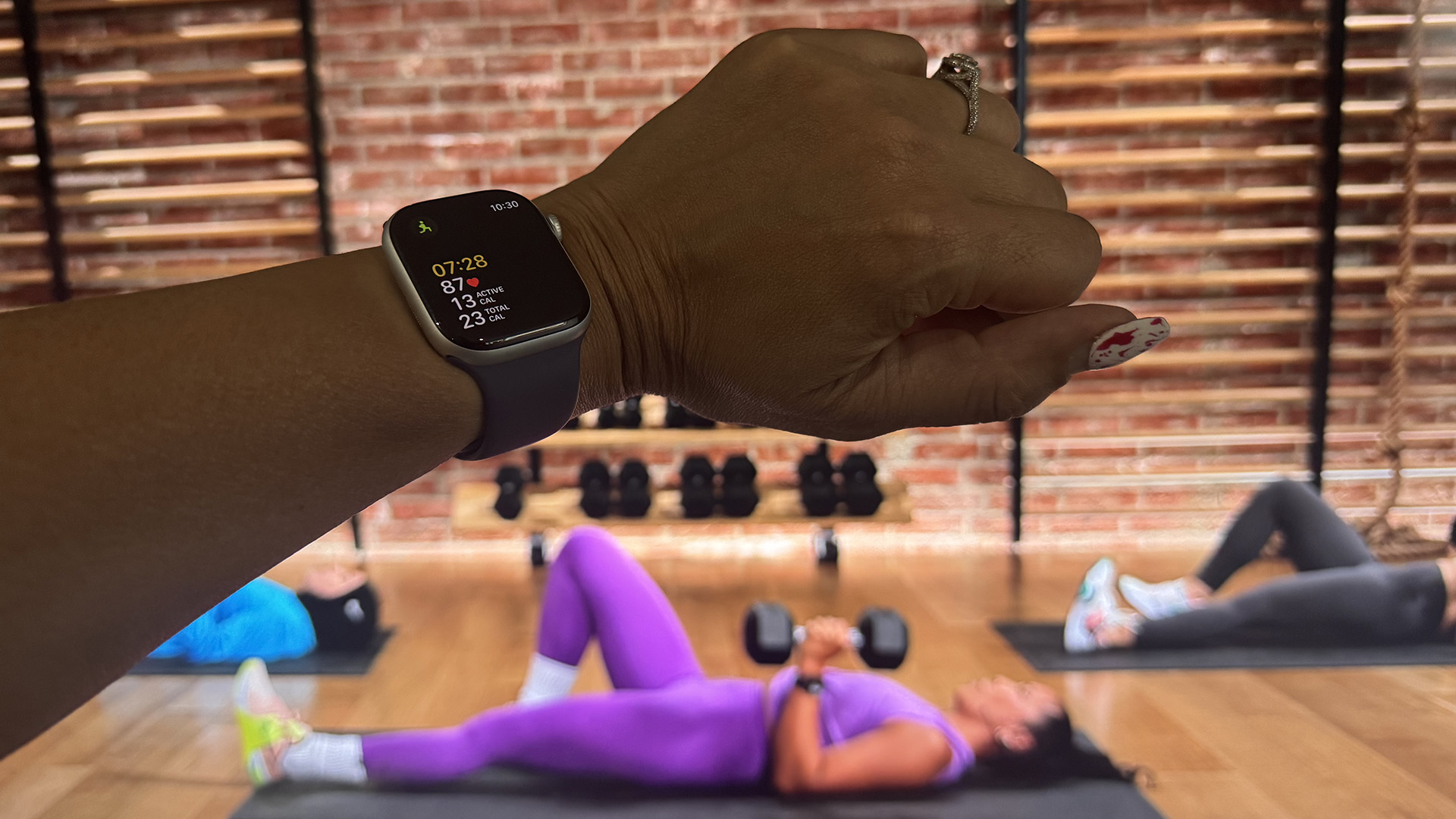 An Apple Watch Series 11 on a wrist tracking a workout with a TV in the background showing Apple Fitness+.