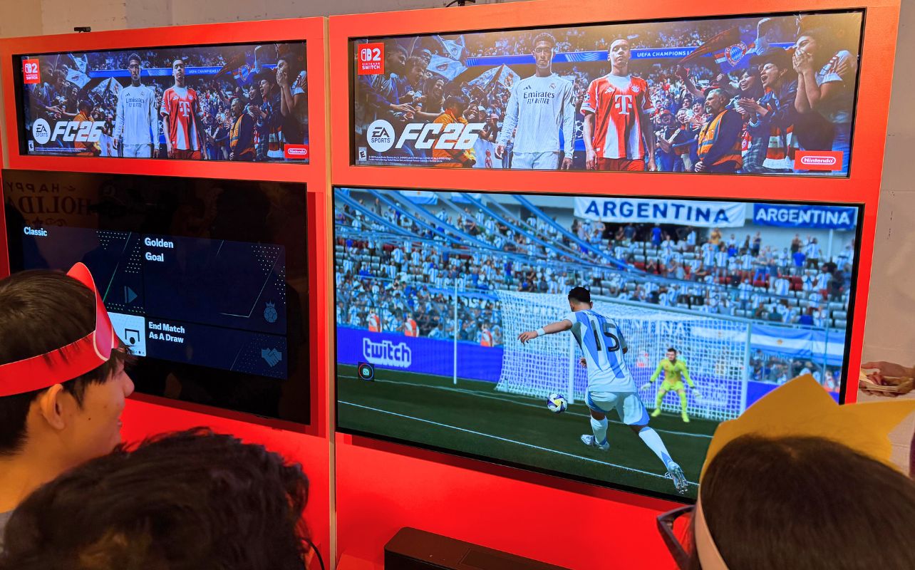 People in crowns watch soccer video game on red displays, showing EA FC 26 .Background has Argentina stadium scene.