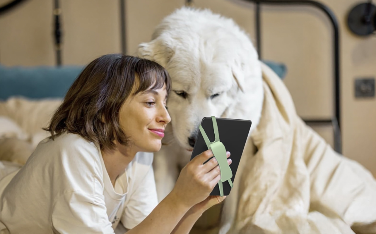 A woman in bed with a strap on an eReader, a dog looking on.