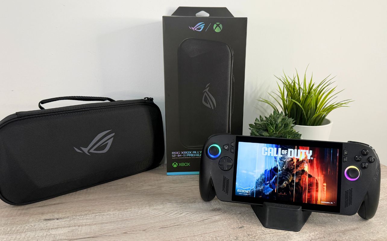 A picture of the ROG Xbox Ally X on a desk with a travel case, the retail box and the ROG Xbox Ally X handheld on a stand.