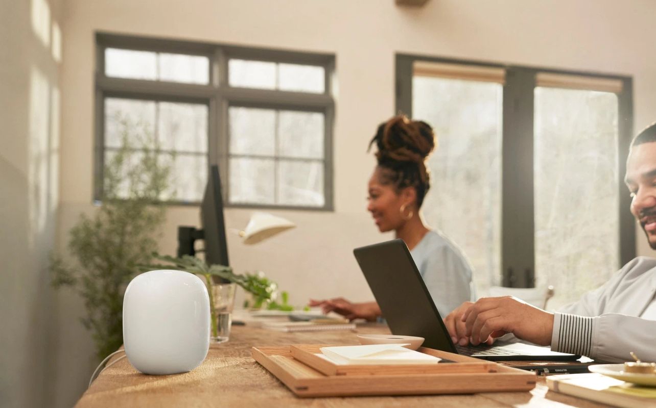 Two people work on computers with a Nest Wi-Fi Pro sitting in the foreground.