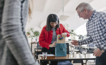 A woman receiving last-minute gifts in a bag from her colleagues and celebrating at the office after a recent success.