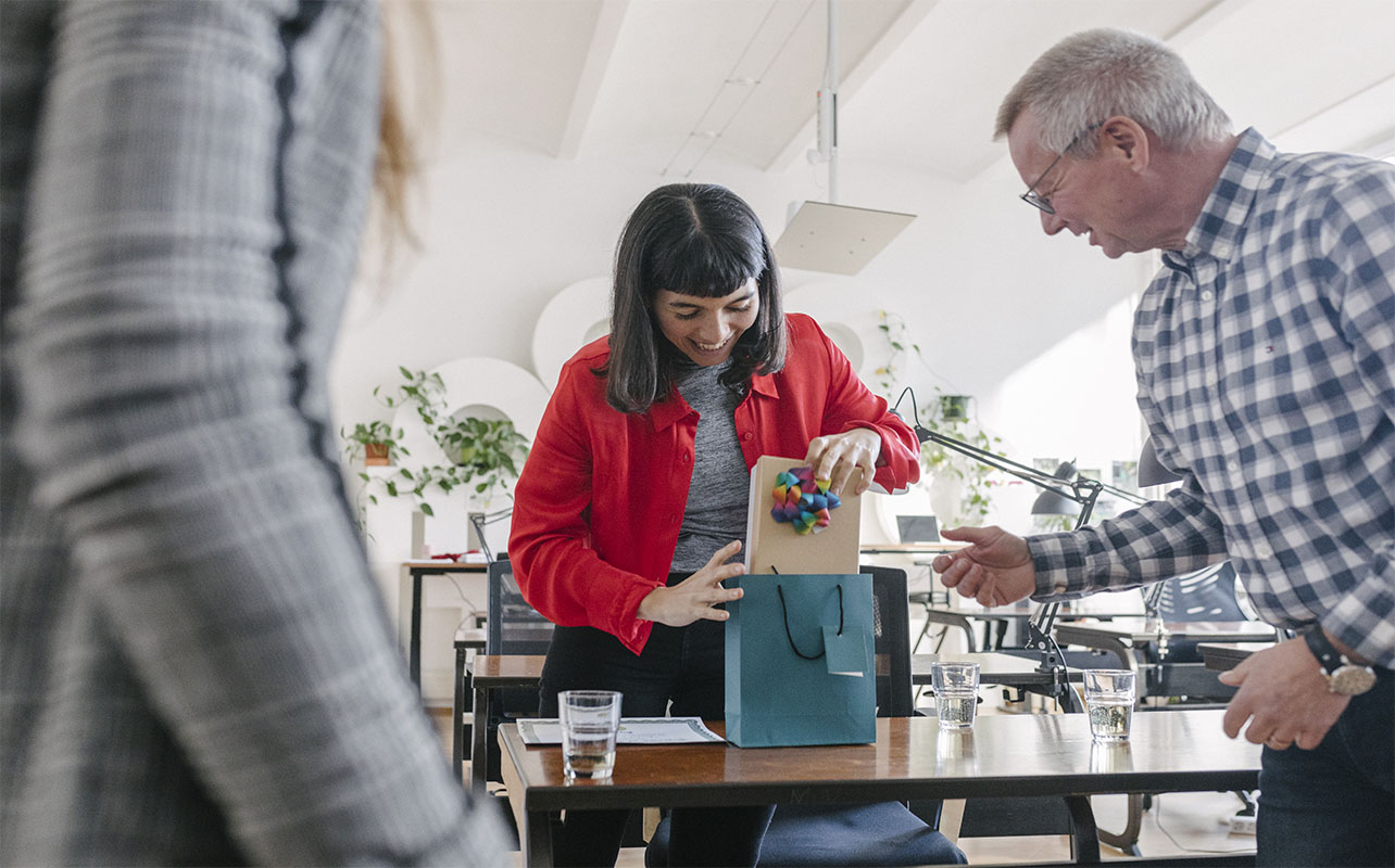 A woman receiving last-minute gifts in a bag from her colleagues and celebrating at the office after a recent success.
