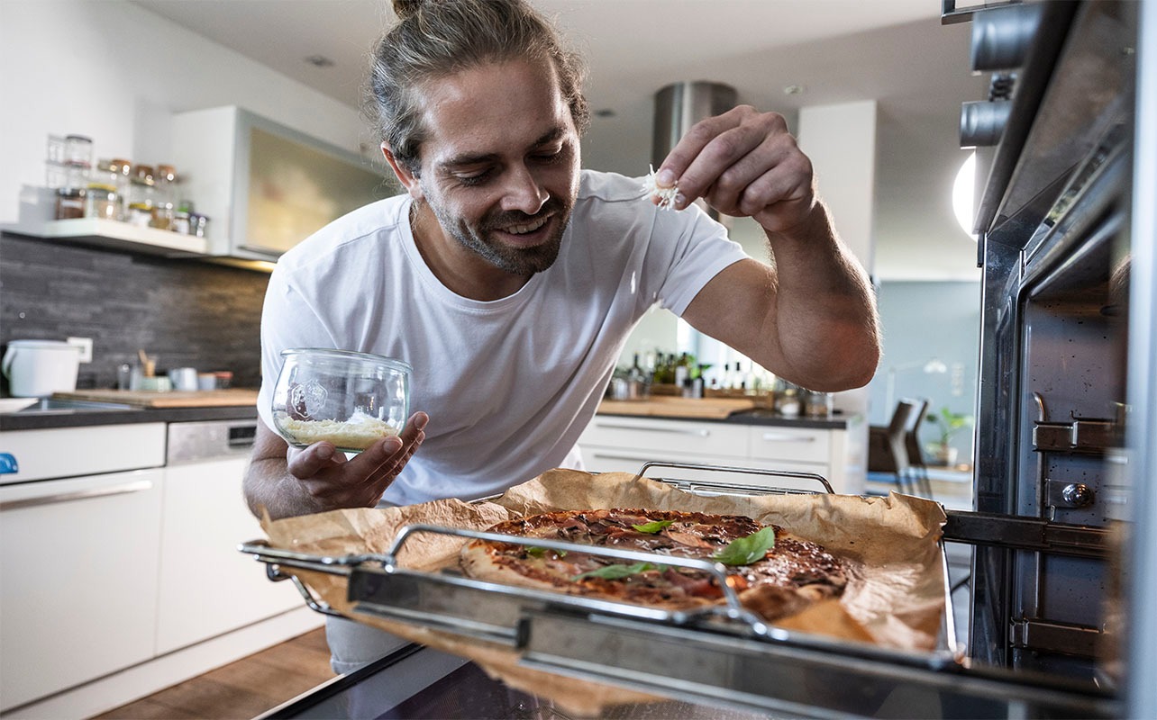 A man adds cheese on a pizza before putting it in a commercial oven.