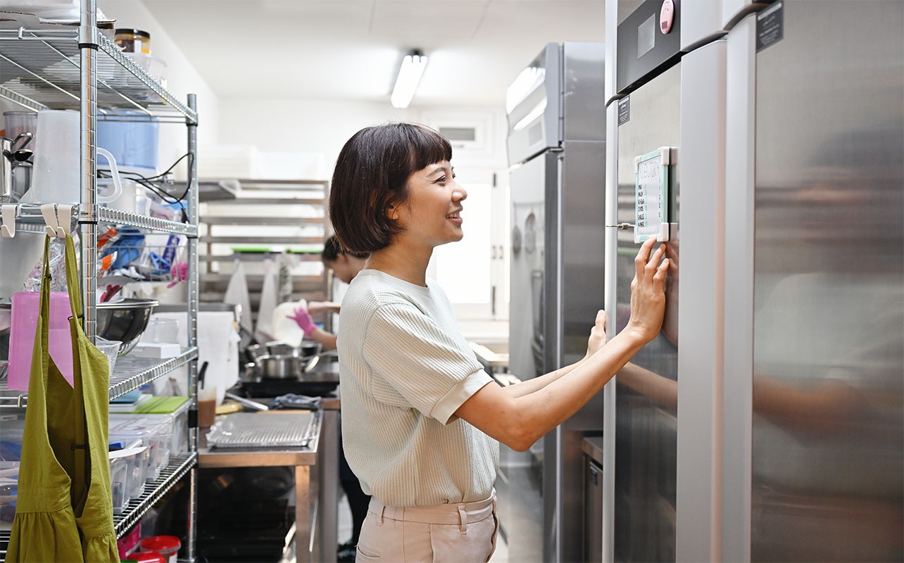 A woman checks notes on a small whiteboard affixed to a commercial refrigerator.