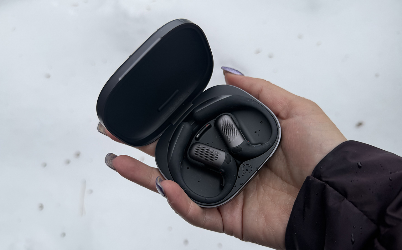 A hand holding the Shokz OpenFit Pro earbuds in front of snow