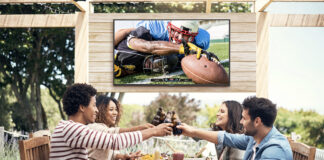 Four friends on a patio clinking glasses, a Neptune Full Sun outdoor TV behind them.
