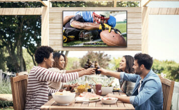 Four friends on a patio clinking glasses, a Neptune Full Sun outdoor TV behind them.