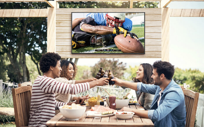 Four friends on a patio clinking glasses, a Neptune Full Sun outdoor TV behind them.