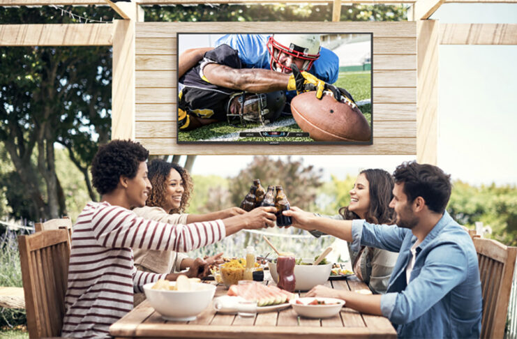 Four friends on a patio clinking glasses, a Neptune Full Sun outdoor TV behind them.
