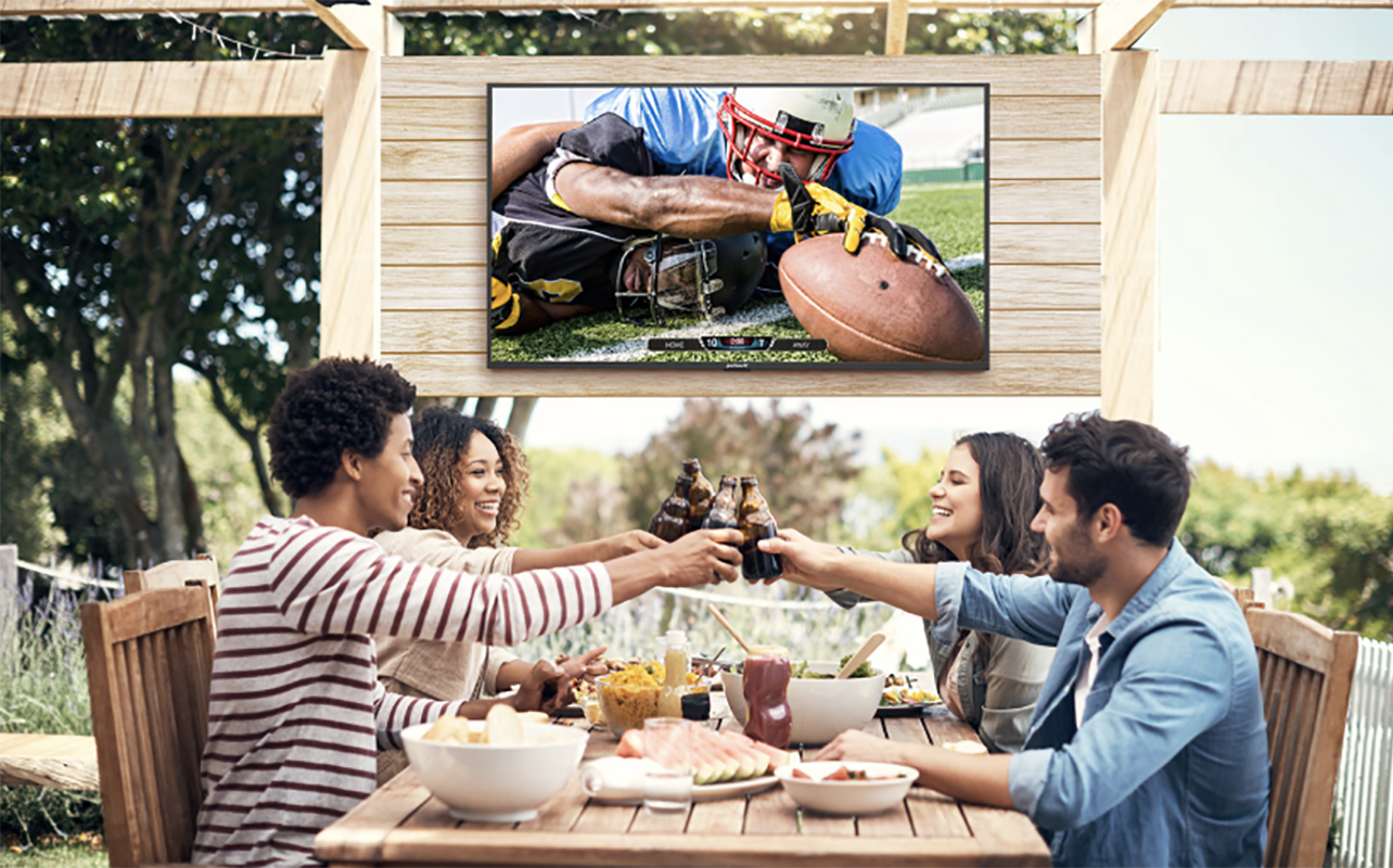 Four friends on a patio clinking glasses, a Neptune Full Sun outdoor TV behind them.