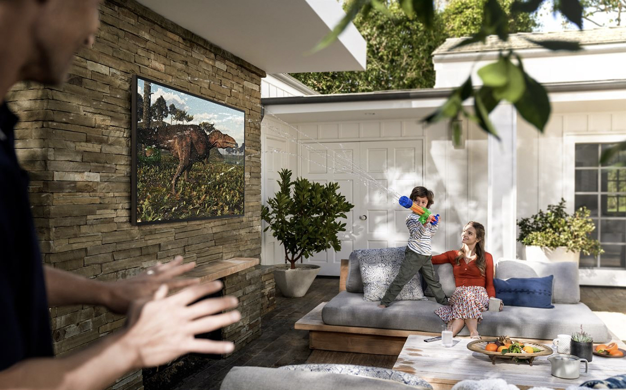 A child spraying a water gun beside a Samsung The Terrace outdoor TV on a deck.