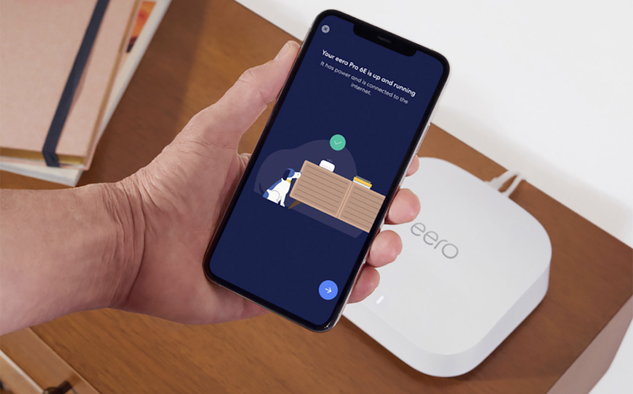A hand holding a phone with the eero app in front of an eero Pro 6 mesh Wi-Fi node.