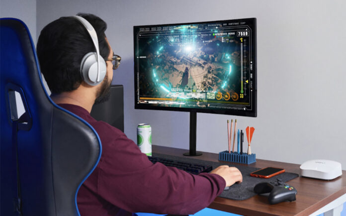 A man sitting at a desk playing games on a computer with an eero Pro 6 mesh Wi-Fi node beside him.