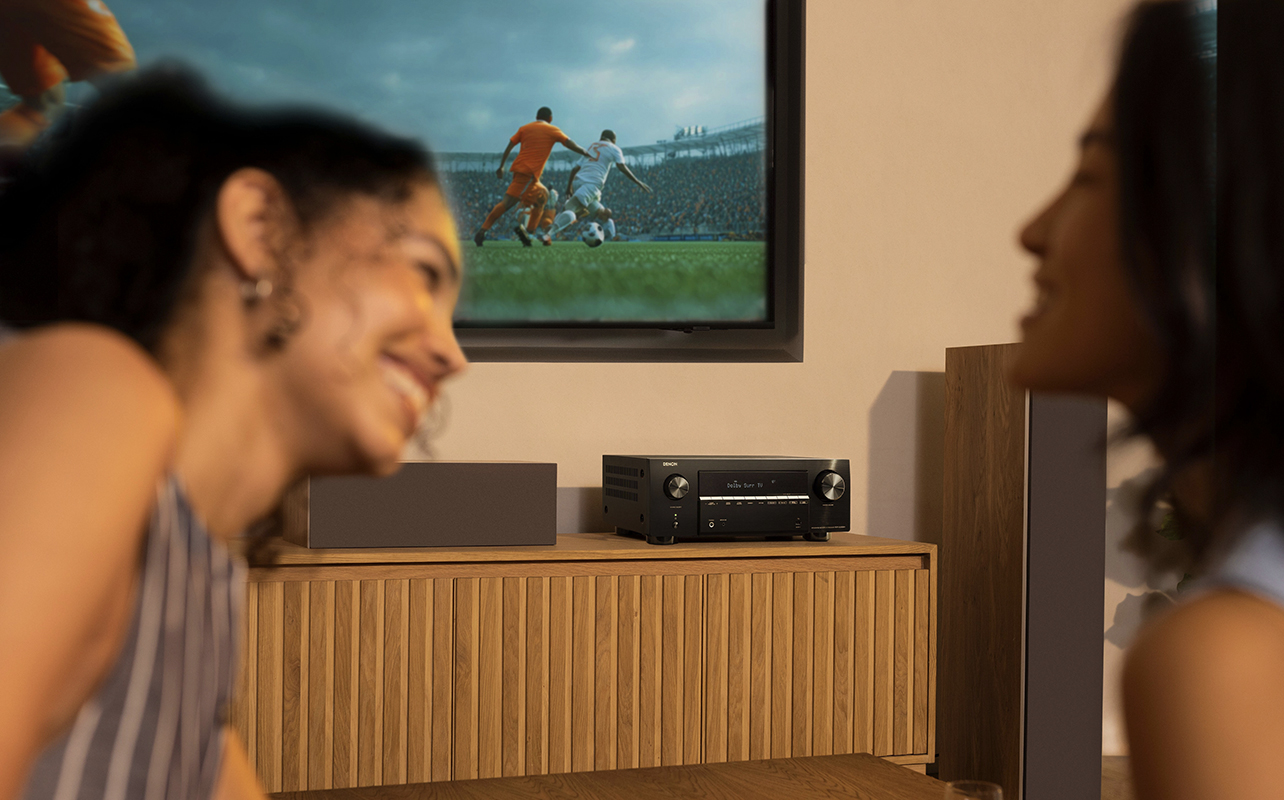 Two women smiling in front of a Denon AVRX2900 AV receiver.
