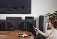A man sitting in the corner of a living room turning on a TV with a Denon AX1700 AV receiver under the display.