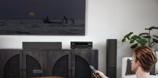 A man sitting in the corner of a living room turning on a TV with a Denon AX1700 AV receiver under the display.