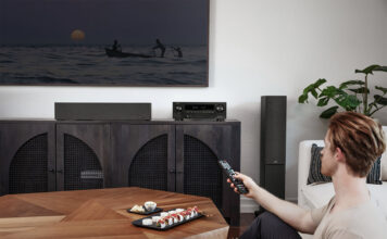 A man sitting in the corner of a living room turning on a TV with a Denon AX1700 AV receiver under the display.