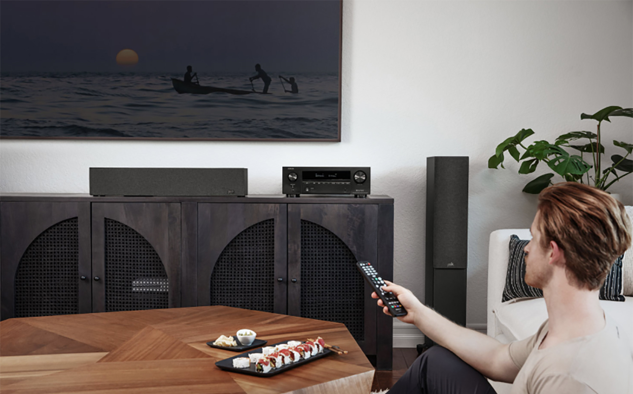 A man sitting in the corner of a living room turning on a TV with a Denon AX1700 AV receiver under the display.