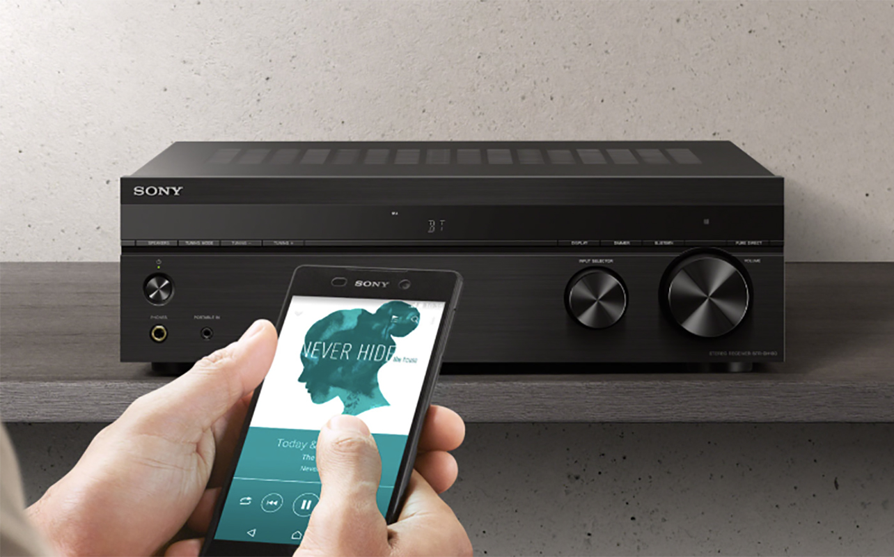 A hand holding a phone with music playing in front of a Sony DH190 AV receiver.