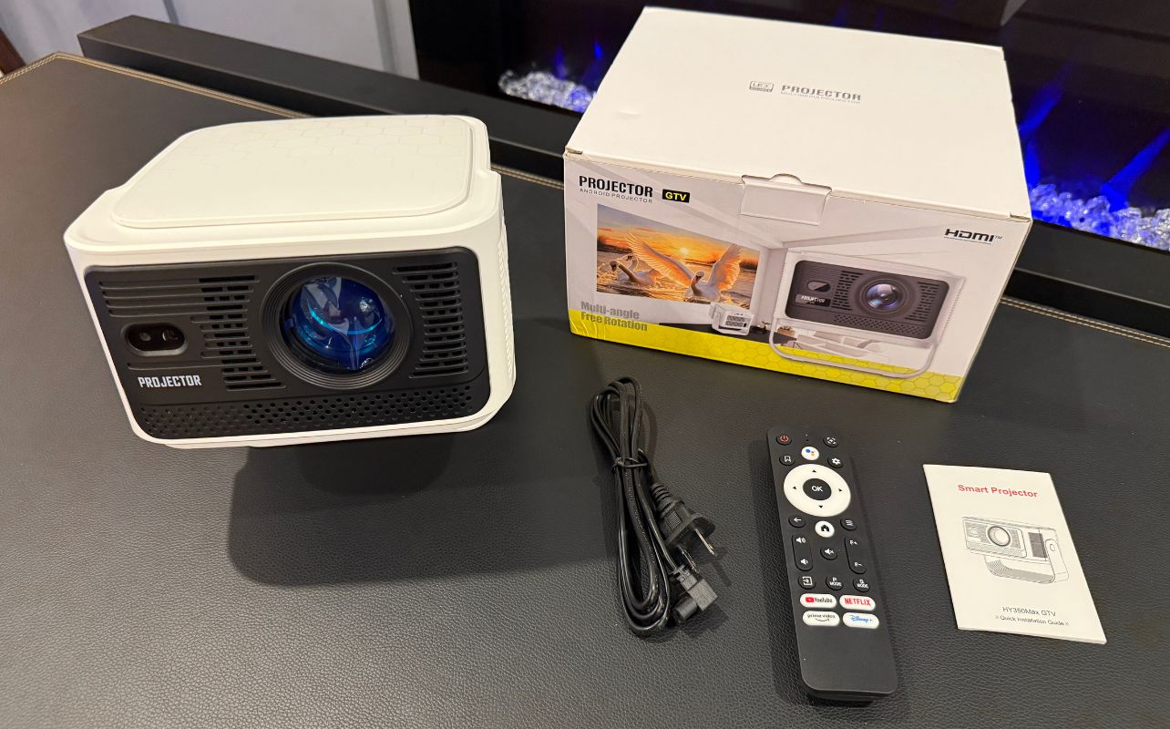 The Brother Eye Mark III Elite Smart LCD Projector is sitting on a desk with all the parts it comes with spread out.