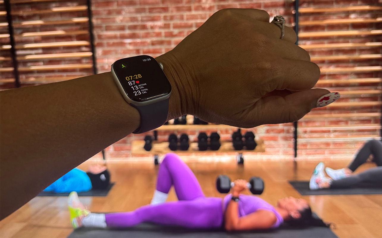 Apple Watch Series 11 on a wrist showing live workout data, the workout on the TV in the background.