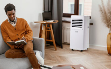 A man sitting on the couch with a Black & Decker portable air conditioner by the window.