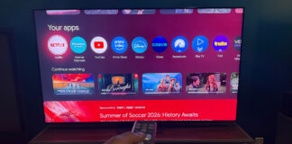 A hand holding the remote in front of the Hisense 65UR9SG RGB mini LED TV.