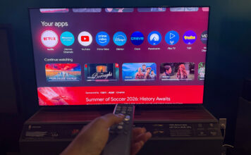 A hand holding the remote in front of the Hisense 65UR9SG RGB mini LED TV.