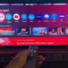 A hand holding the remote in front of the Hisense 65UR9SG RGB mini LED TV.