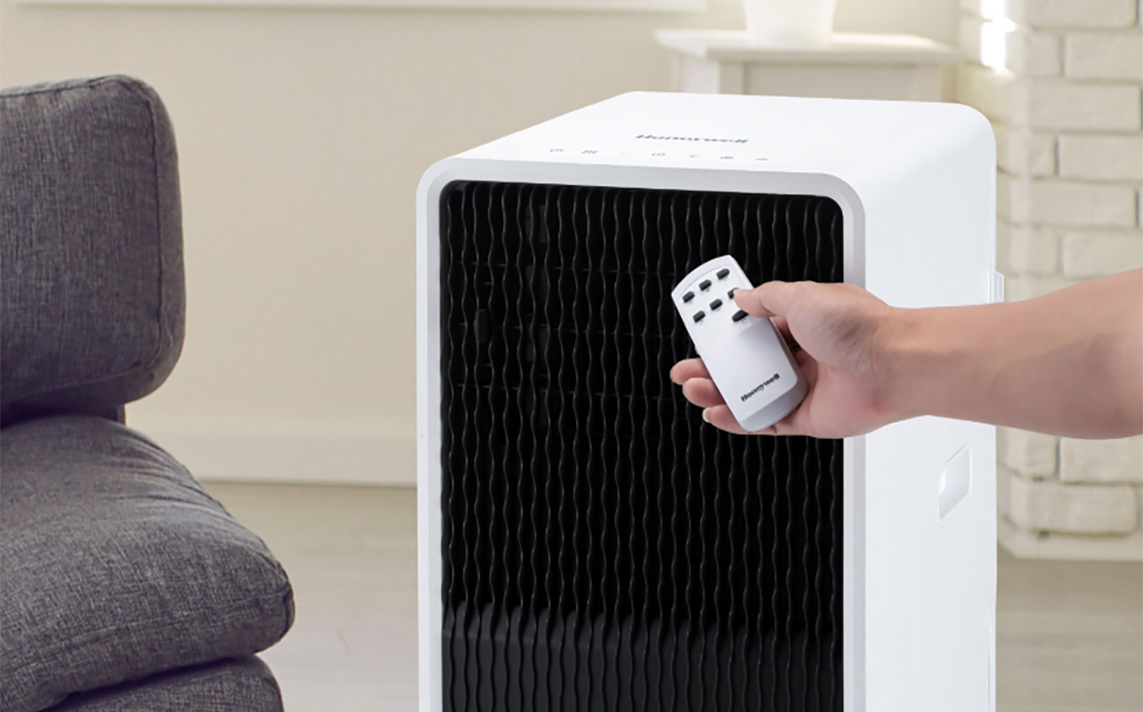 A hand holding a remote beside a Honeywell 3-in-1 portable air conditioner.