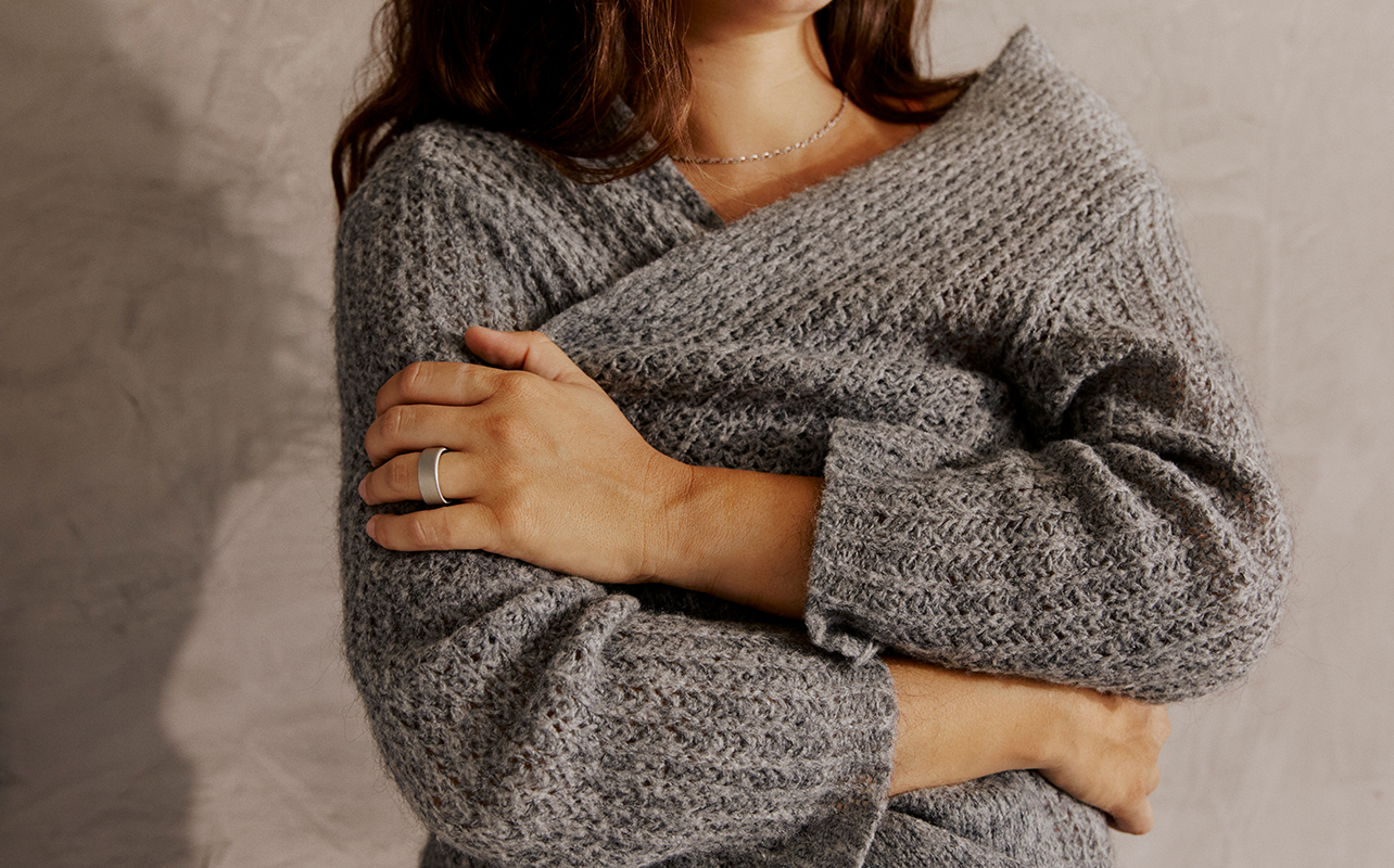A woman wearing a fuzzy sweater hugging herself with an Oura Ring 5 smart ring on her finger.