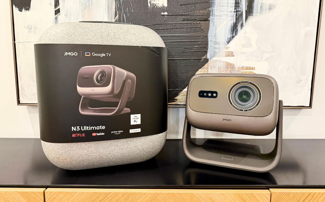 A retail box and the JMGO N3 Ultimate projector are sitting next to each other on a black tabletop with a painting in the background.