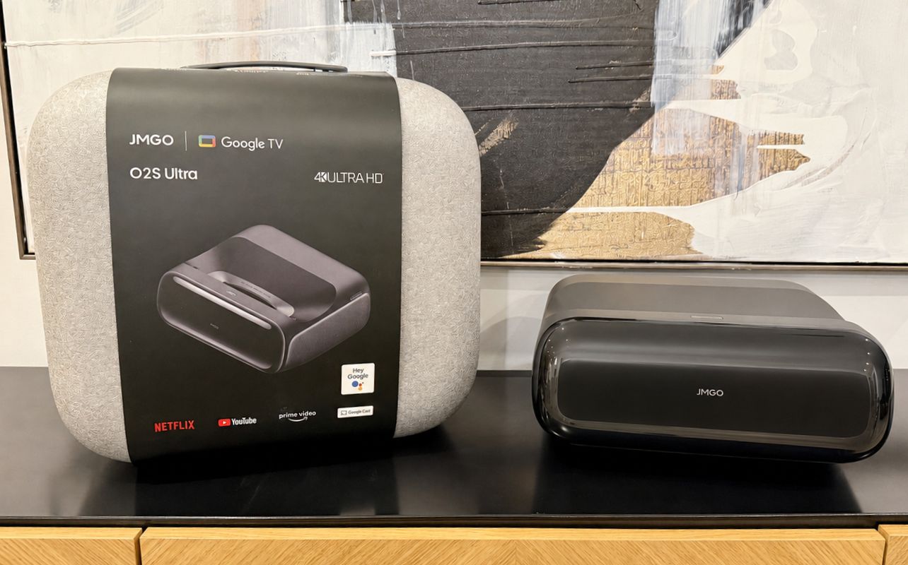 A retail box and the JMGO projector are displayed on a black tabletop with a painting in the background.