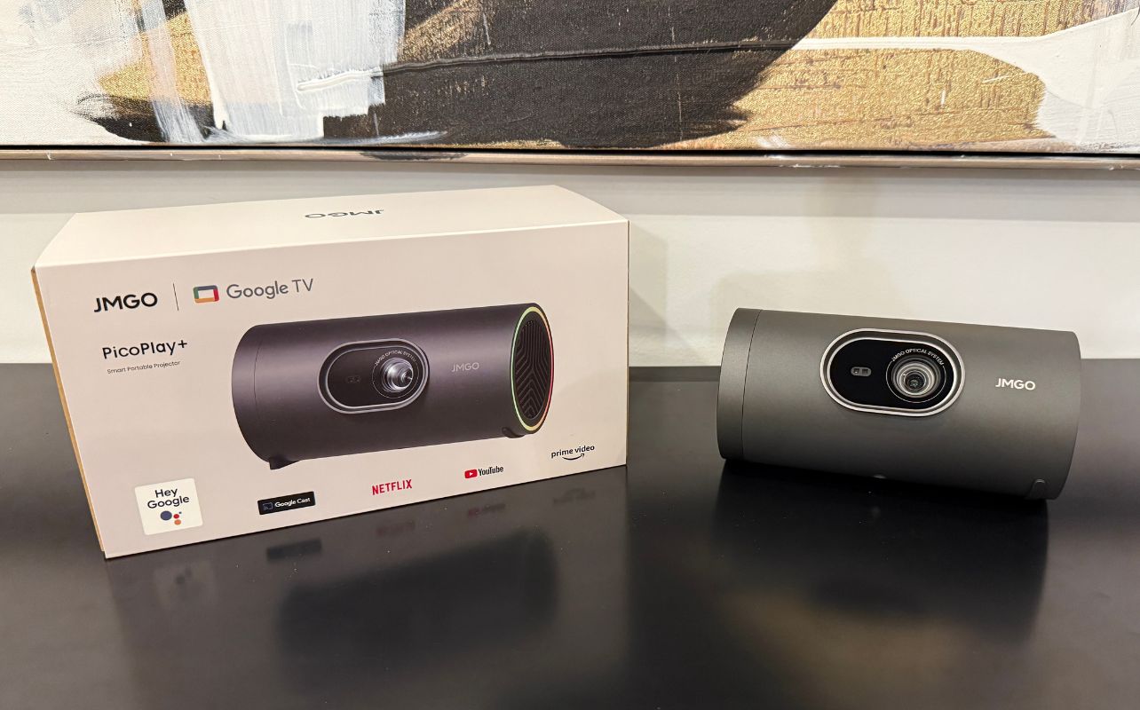 A retail box and the JMGO PicoPlay+ projector are displayed on a black tabletop with a painting in the background.