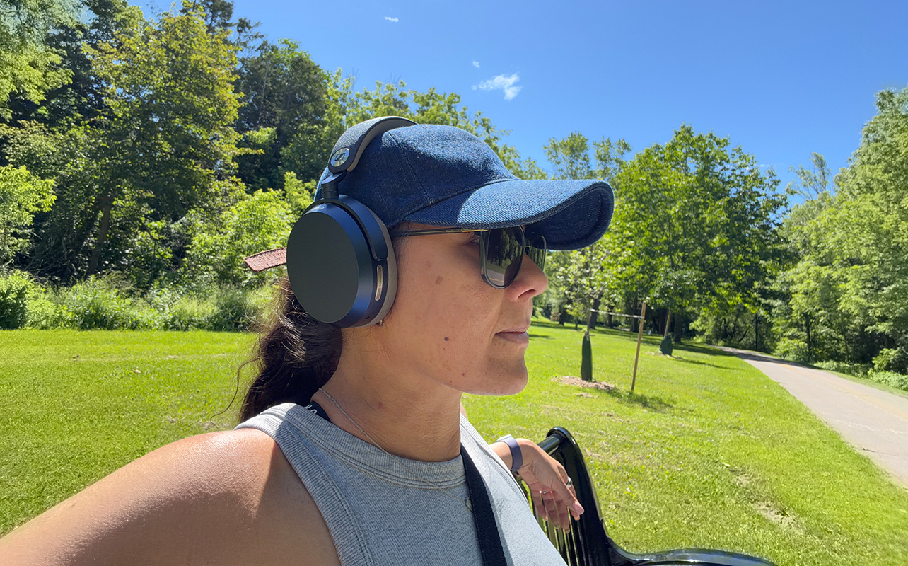 A woman sitting on a bench outdoors wearing the Sennheiser Momentum 5 Wireless headphones.