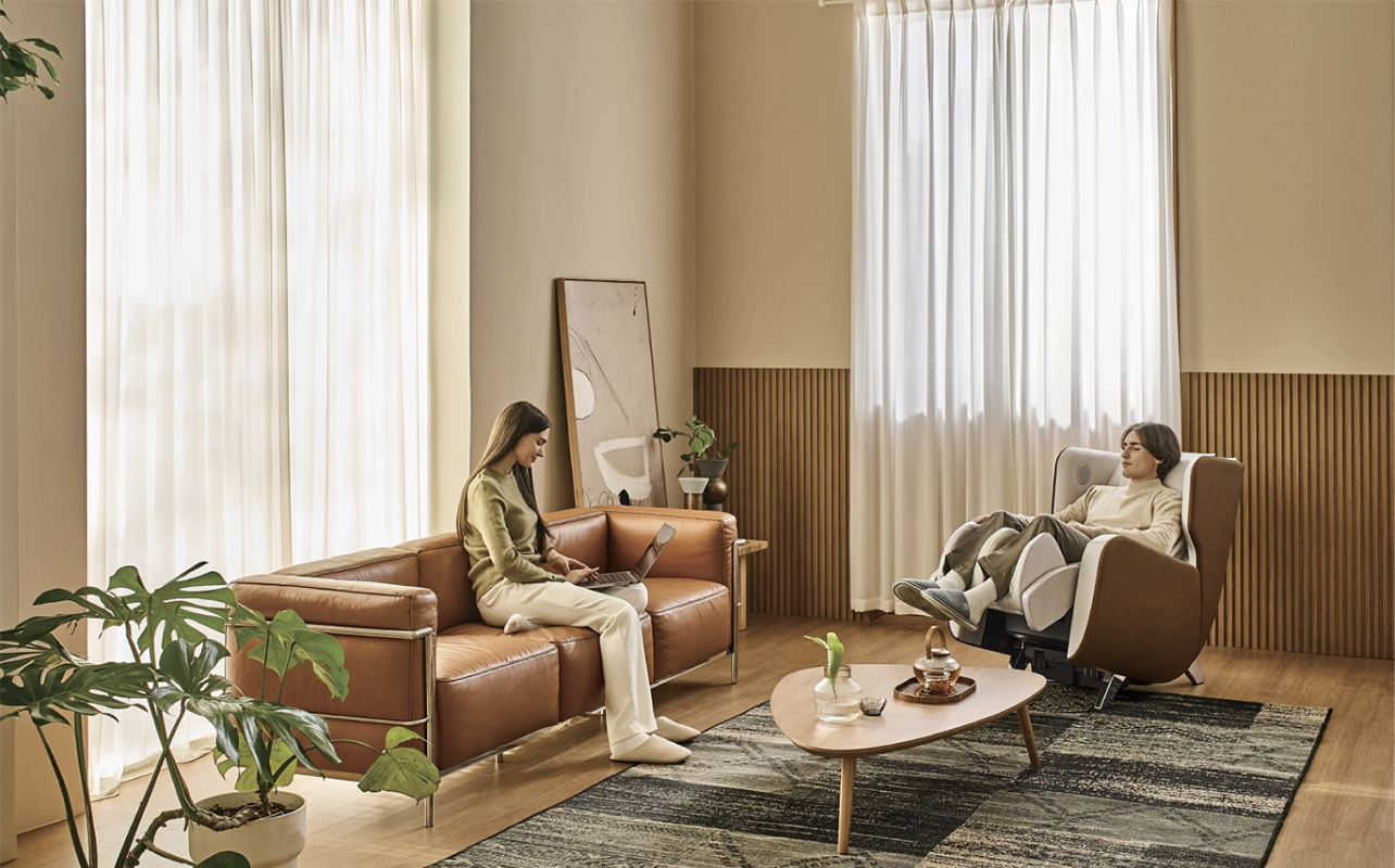 A woman reading a book in a living room while a man lies back in a Bodyfriend Eliza massage chair.