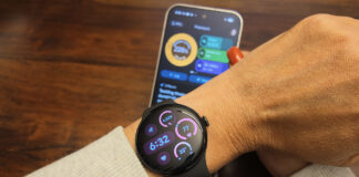 The Google Pixel Watch 5 on an arm with the Google Pixel 11 Pro in the background showing the Google Health app