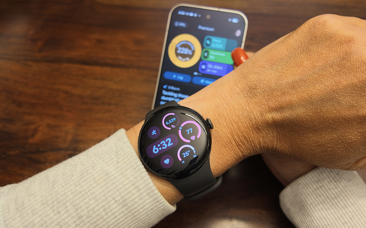 The Google Pixel Watch 5 on an arm with the Google Pixel 11 Pro in the background showing the Google Health app