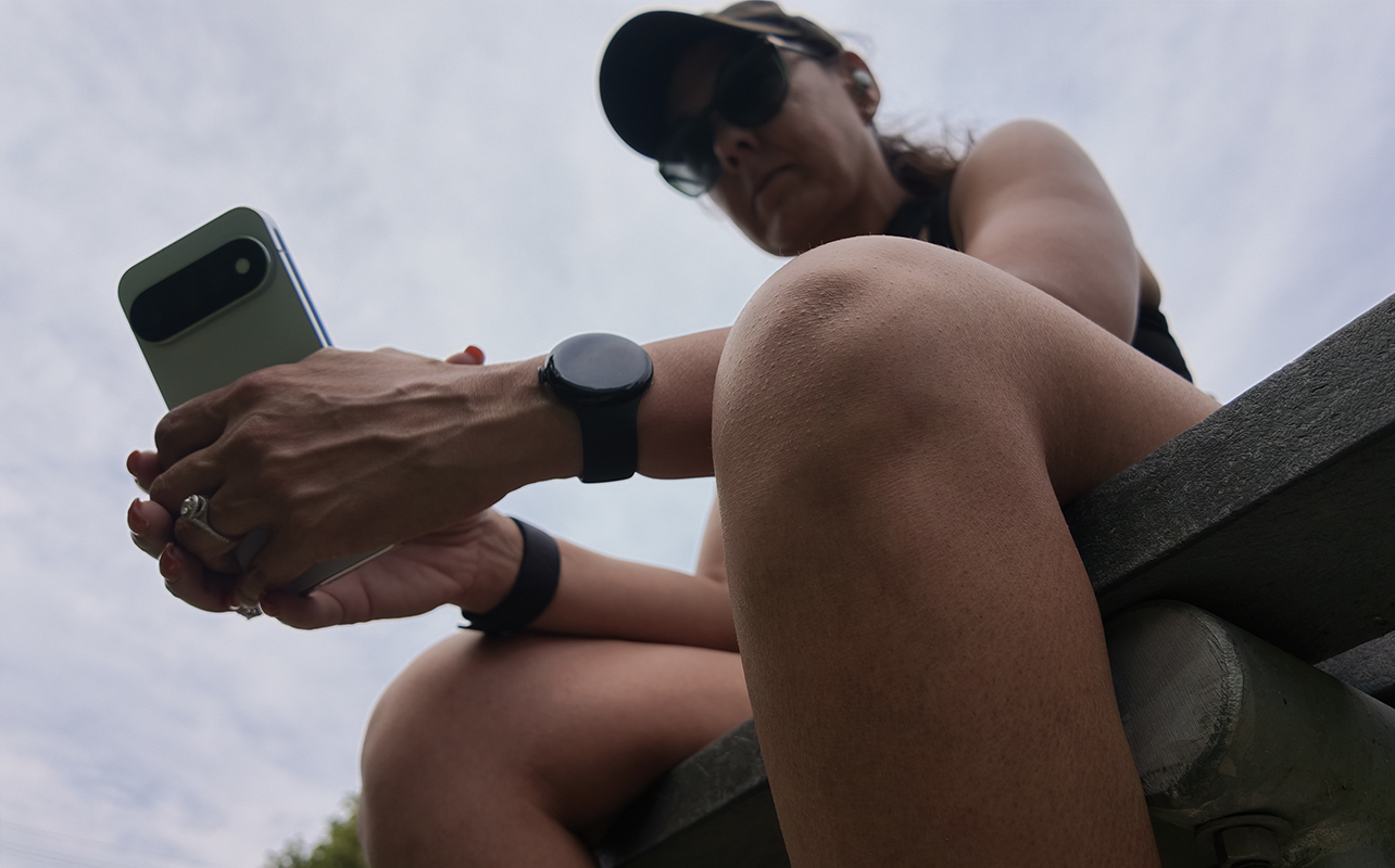 An underhand shot of a woman sitting on a bench outside holding a Google Pixel 11 Pro phone and wearing the Google Pixel Watch 5.